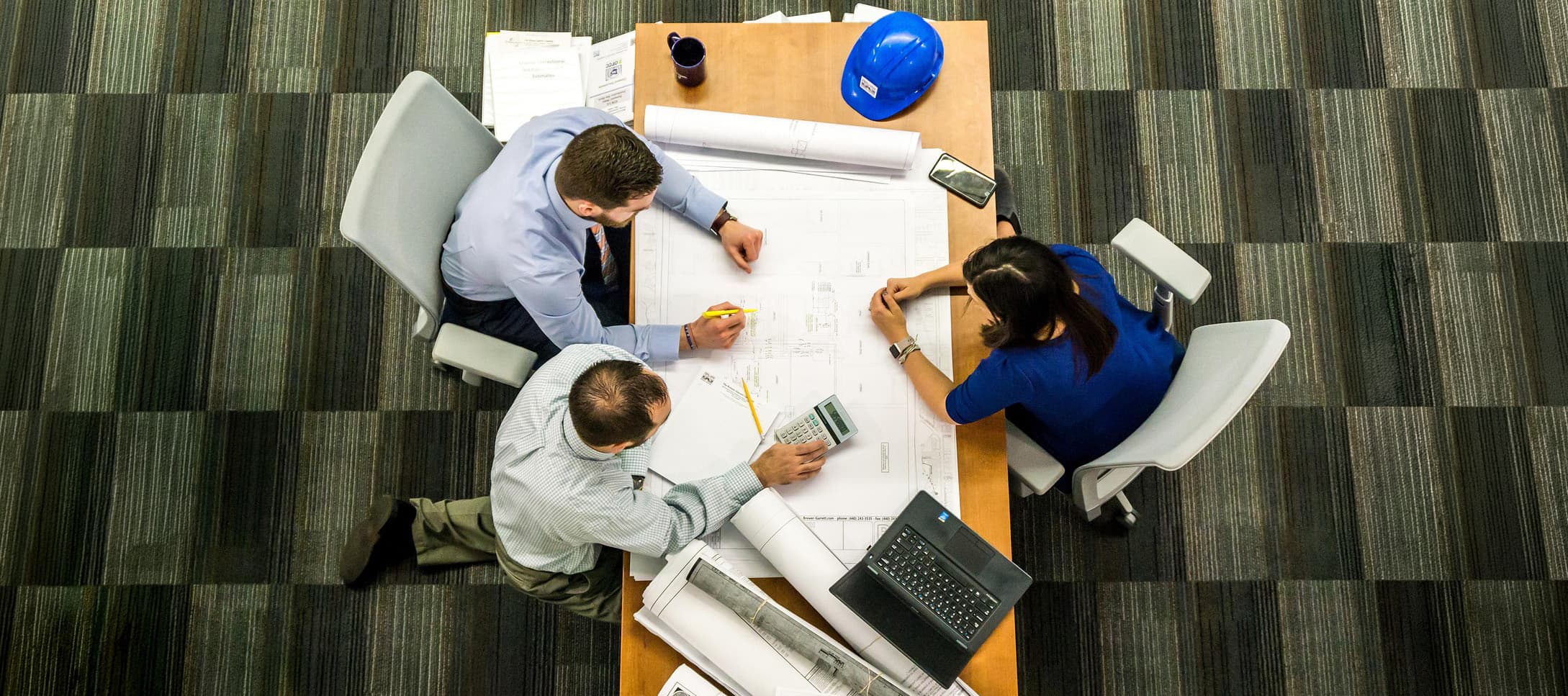 Three professionals collaborating over architectural blueprints and plans with a helmet and laptop on the table.